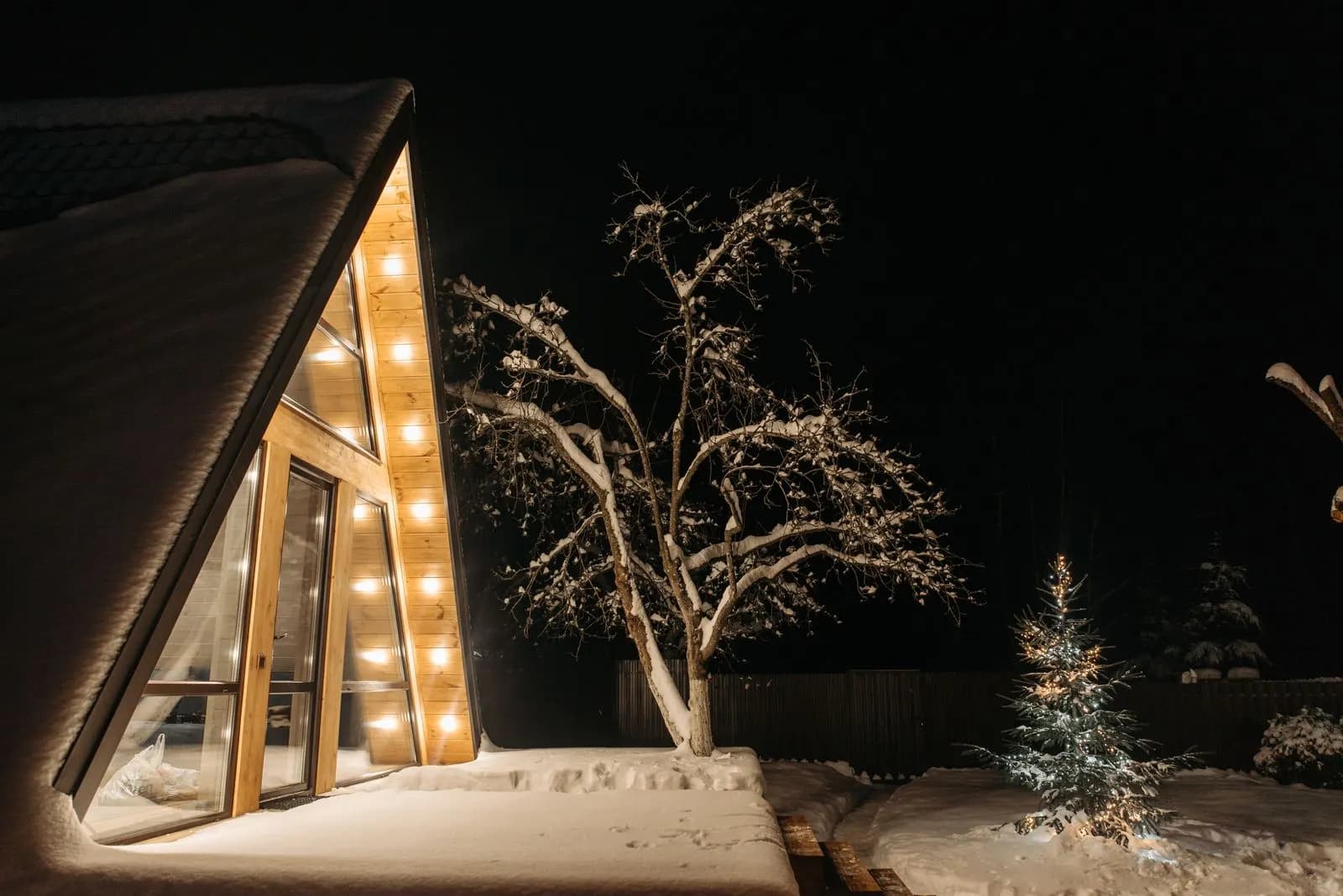 Example of a modern A-frame home with warm white bulbs along the gable and a lit evergreen in the yard