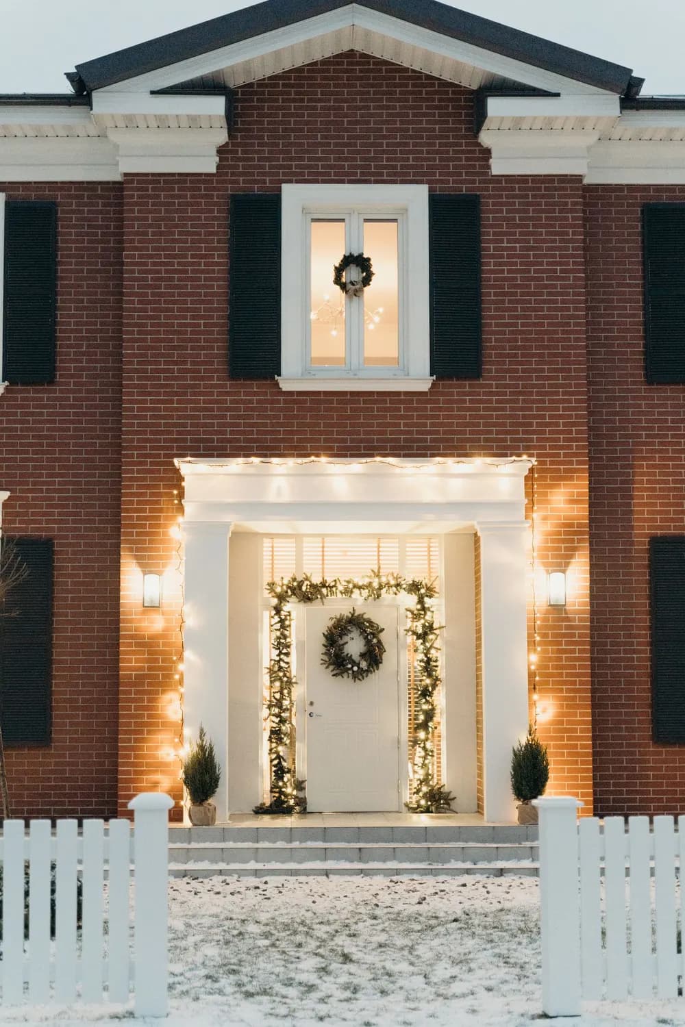 Example of a brick home with warm white lights framing the front entry, a lit garland and a wreath
